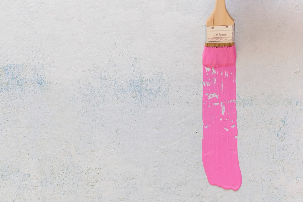 A paintbrush applies a vibrant pink streak on a textured white wall, conveying creativity and spontaneity.