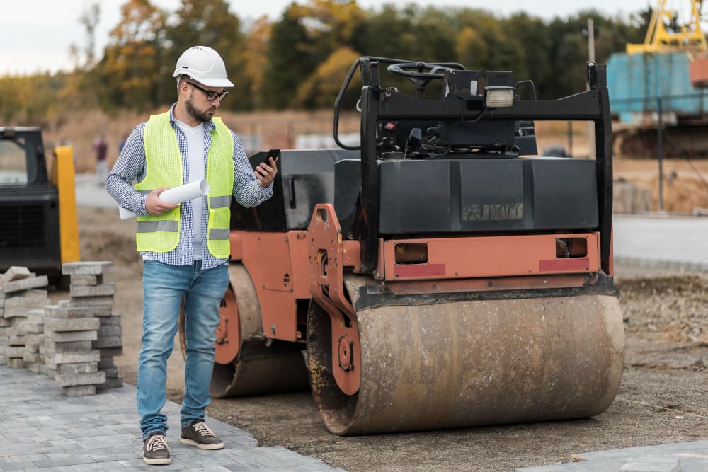Man in a hard hat and neon vest checks phone, holding blueprints near a steamroller on a construction site.