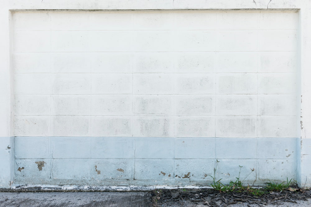 Worn white brick wall with blue stripe, peeling paint, and dirt; sparse grass and concrete add texture.