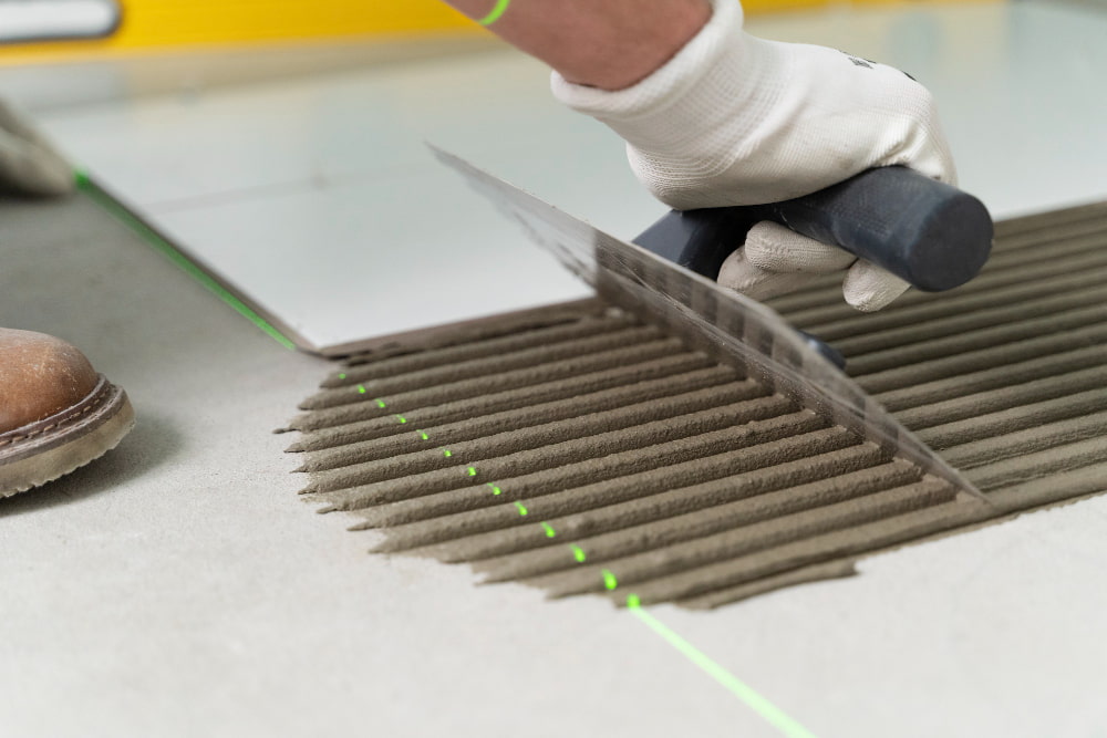 Gloved hand spreads tile adhesive with notched trowel; green laser line guides precise floor alignment.