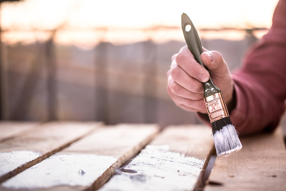 A hand in a red sleeve holds a paintbrush, applying white paint to wooden boards.
