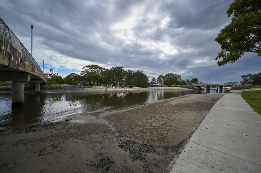 Overcast sky above a calm river with two bridges. People walk on a sandy shore, surrounded by lush trees.