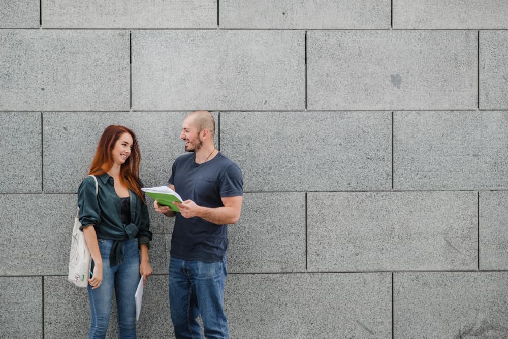 Smiling man and woman in jeans chat by a gray stone wall; man holds a notebook, both appear engaged.